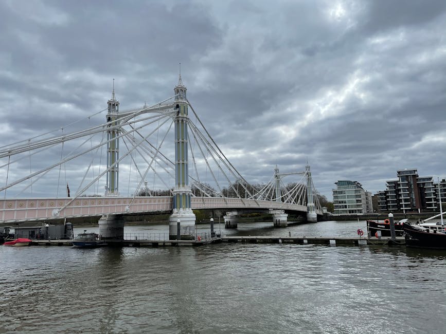 View of the Hammersmith Bridge spanning the River Thames under a cloudy sky, with its suspension cables and ornate towers visible. In the foreground, there is a small pier with several boats docked, and part of a modern residential building can be seen on the right side. The scene indicates an urban riverside environment typical for London. Although not directly related to moving services, this image provides context for the area's architecture and scenery relevant to house relocations near Hammersmith. Man and Van Hammersmith facilitates such relocation processes, including packing and furniture transport, in this scenic district.