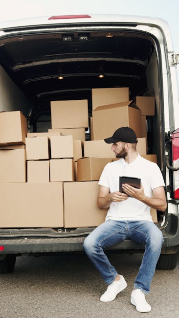 A man wearing a white t-shirt, blue jeans, white sneakers, and a black cap is sitting on the edge of the open back of a white moving van, which is loaded with stacked cardboard boxes of various sizes. The boxes are secured with plastic wrapping and occupy most of the van’s interior space, situated on a paved surface. The man is holding a tablet and appears to be reviewing information related to a home relocation or packing process. The scene is outdoors with natural daylight, and the background features a neutral setting suitable for household removals, illustrating the packing and loading stage of a furniture transport or house move, aligned with services offered by Man and Van Hammersmith.
