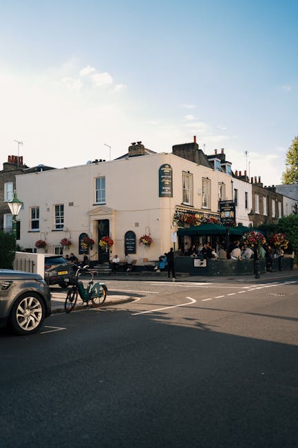 A street view of a white, three-storey building located on Hammersmith Broadway, with a pub or restaurant on the ground floor featuring a dark green awning and hanging flower baskets. The building has multiple windows, some with flower boxes, and a sign indicating it offers food and drink. In front of the building, several people are seated outside at tables, enjoying the outdoor seating area. Parked vehicles, including a black car and a bicycle secured to a bike rack, are visible on the pavement. The scene is illuminated by daylight with a clear sky overhead, and the environment suggests a busy, urban setting suitable for house relocations or furniture transport supporting services like those offered by Man and Van Hammersmith.