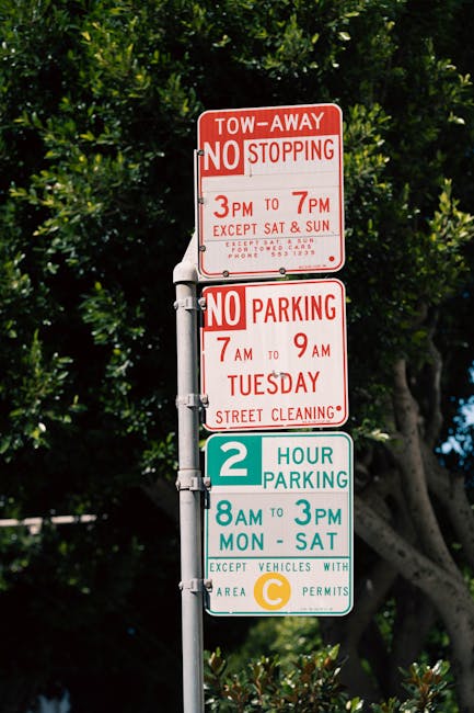 A tall metal parking regulation post featuring multiple signs is set against a background of green trees with dense foliage. The topmost sign is red and white, indicating a tow-away zone with no stopp