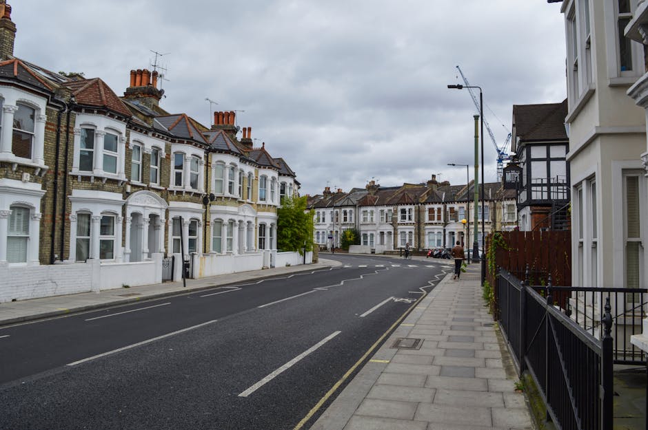 A quiet residential street in Hammersmith showing a row of Victorian-style terraced houses with white facades, bay windows, and red-tiled roofs. The pavement on the right side is lined with black metal railings and a few potted plants. The street appears temporarily clear of traffic, with a few parked cars visible further down. Overhead, the sky is overcast with grey clouds, creating a subdued lighting environment. This setting represents an area where home relocation and furniture transport services by Man and Van Hammersmith are commonly performed, with the street providing access for loading and unloading furniture and boxes near the accessible entrances of the houses.