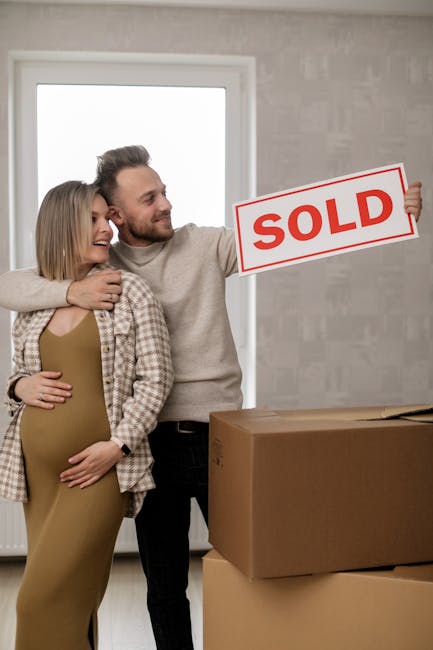 A couple stands inside a modern property with a large window and neutral grey walls, smiling and celebrating a house sale. The man is holding a white and red 'SOLD' sign with both hands, positioned to the right side of the image. The woman, visibly pregnant, is standing close to him with her arms around his waist. In front of them on the floor is a large, flat cardboard box, possibly for packing or moving purposes. Natural light enters through the window, illuminating the scene, which captures a moment of achievement related to home relocation or real estate transaction. This setting, combined with the cheerful expressions, aligns with themes of house moves and property transactions, relevant to a removals service context at manandvanhammersmith.org.