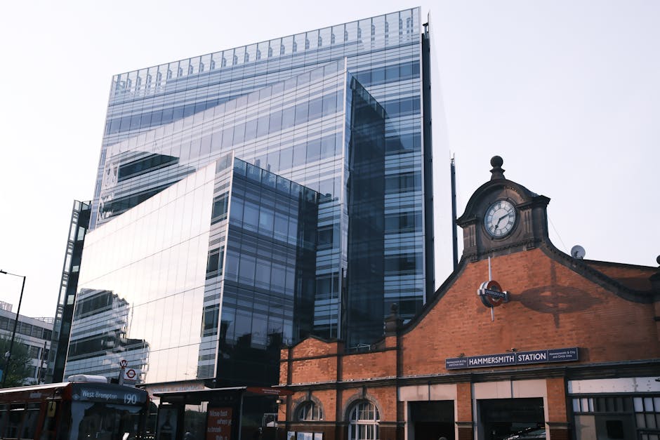 A modern, tall office building with a glass facade reflecting the surrounding cityscape, situated behind the historic brick structure of Hammersmith station entrance, which features a clock and a sign indicating the station's name. In front of the station, a bus with a route number 190 is visible, parked on the street pavement. The image is taken during daylight with clear, bright lighting, highlighting the contrast between the contemporary building materials and the traditional station architecture. The scene captures elements relevant to house removals and furniture transport, illustrating urban moving logistics near the W6 postcode area, with [COMPANY_NAME] providing professional removals services in a busy London environment.