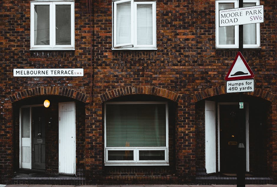 A brick residential building on Melbourne Terrace in Hammersmith, displaying two white-framed windows on the upper floor and a ground-floor window with closed blinds. The building has an arched brick entrance with a black door and a white wooden door on the left side, illuminated by a small external wall light. The exterior includes two street signs, one indicating Melbourne Terrace SW6 and another with Moore Park Road SW6, along with a road warning sign about upcoming bumps for the next 450 yards, attached to a black pole. The scene is captured in natural light, with no visible moving or packing activity, though it represents the typical environment where [COMPANY_NAME] may perform house removals, furniture transport, or home relocation services within this area.